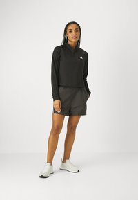 Maglietta nera con zip e maniche lunghe abbinata a pantaloni corti scuri e scarpe da ginnastica bianche. Branding evidente sulla maglietta e sui pantaloni.
