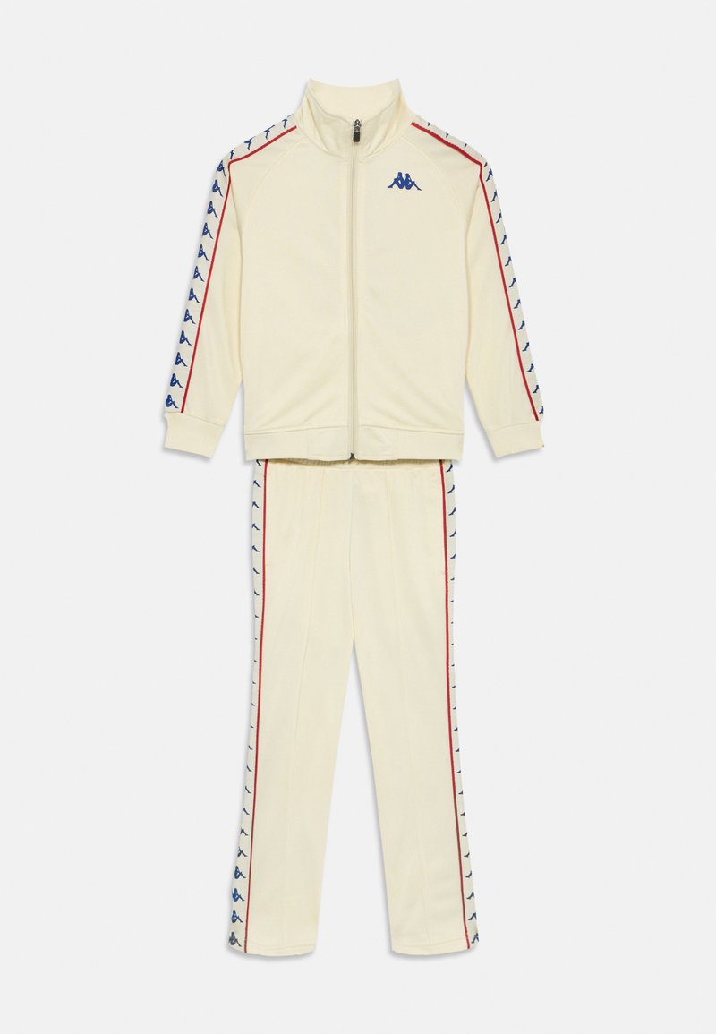 Kremni trenirka iz mehkega materiala. Ima jopico z zadrgo z rdečimi in modrimi detajli ter ujemajoče se hlače z stranskimi črtami in logotipom.