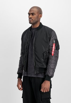 Uomo in piedi che indossa una giacca bomber nera con un'etichetta rossa "Rimuovere Prima del Volo" sulla manica sinistra e pantaloni cargo neri.