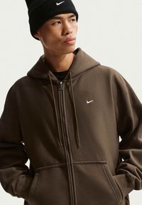 Joven con un gorro negro de Nike y una sudadera con cremallera marrón con el logo blanco de Nike, mirando hacia un lado con las manos en los bolsillos.