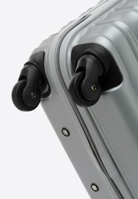 Valigia rigida argentata con superficie testurizzata, due ruote girevoli nere e una chiusura a zip con rivetti in metallo per una maggiore durata.