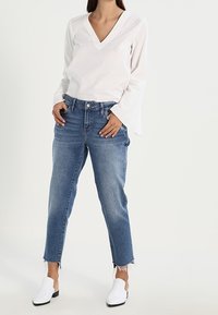 Blouse blanche à manches longues avec un col en V et des poignets évasés, associée à un jean taille haute bleu clair avec un ourlet brut. Chaussures blanches à enfiler.