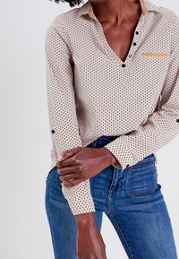 Blouse à manches longues avec un motif à pois beige et bleu marine, un col, des boutons noirs et une poche poitrine sur un tissu doux et texturé.