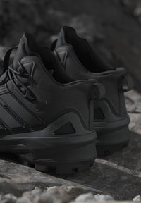 Een paar zwarte sportschoenen met gestructureerde zolen die rusten op een rotsachtige ondergrond in een buitenomgeving.