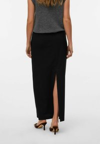 Jupe maxi noire côtelée avec fente sur le côté, associée à un haut sans manches gris. Mannequin portant des chaussures à talons bruns, avec de longs cheveux ondulés.