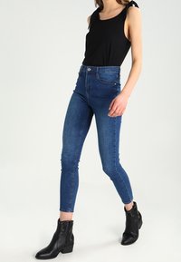 Vrouw die een zwart mouwloos topje met schouderbandjes, hoge blauwe skinny jeans en zwarte enkelboots met studs draagt, die naar voren loopt.