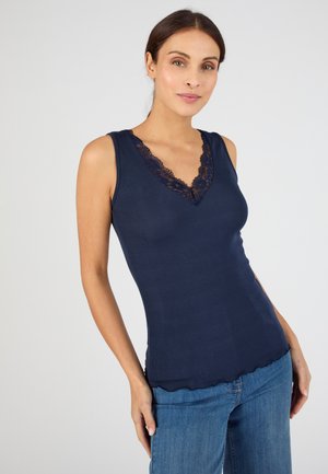 Donna con top senza maniche blu navy con scollo a V in pizzo e jeans blu, in piedi davanti a uno sfondo chiaro uniforme.