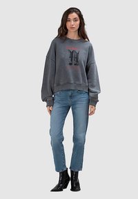 Grijze, oversized sweater met een grafisch ontwerp met zwarte laarzen en rode tekst, gecombineerd met lichtblauwe spijkerbroek en zwarte enkellaarsjes.