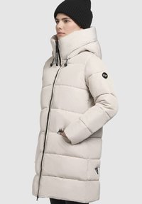 Beige pufferjack met een hoge kraag, verticale gestikte patroon, zijzakken en een logo-opdruk op de linker mouw. Ritsluiting en verstelbare capuchon.