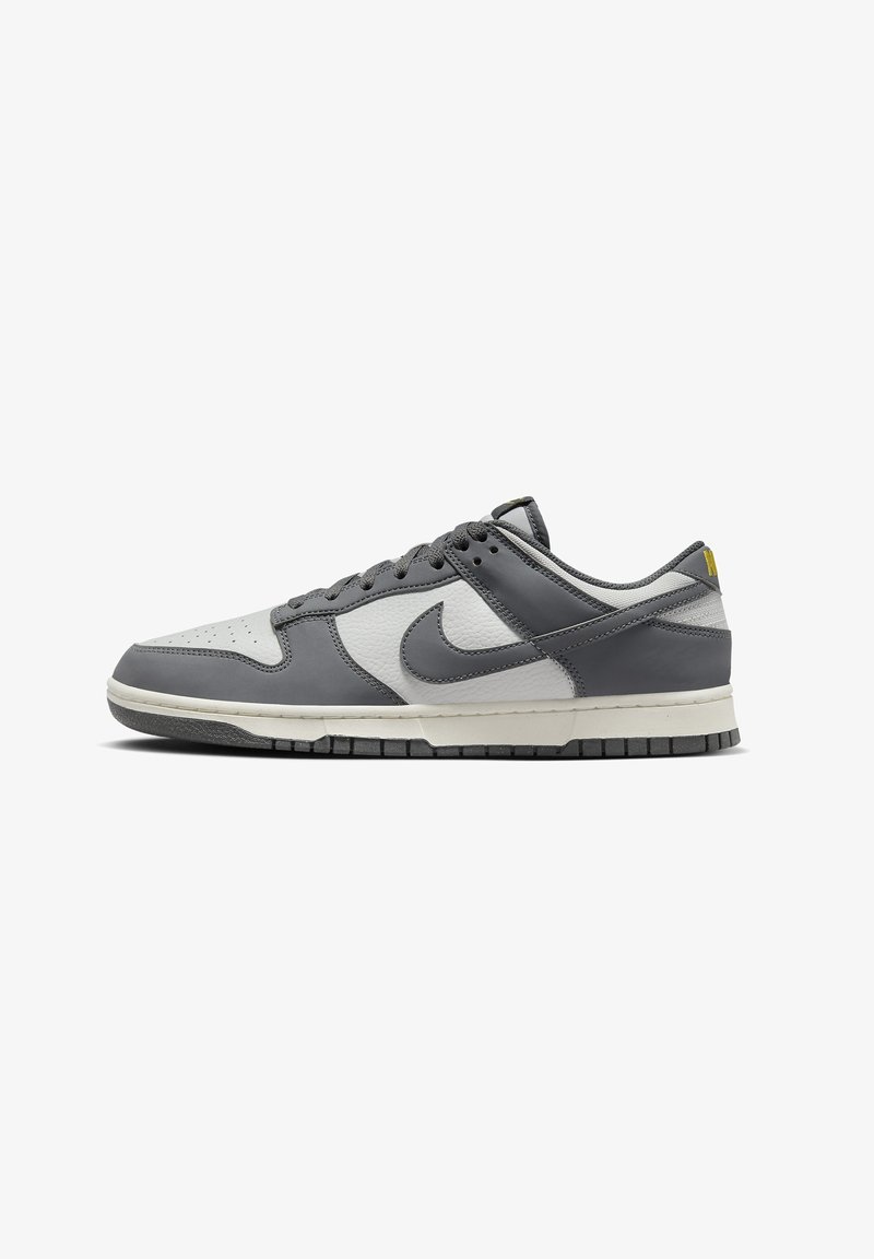 Šedé a bílé kožené tenisky Nike Dunk mají texturovaný svršek, polstrovaný límec a černou gumovou podrážku se žlutým akcentem na patě.
