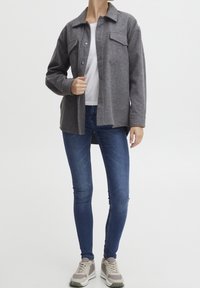 Szara kurtka z mieszanki wełny z kołnierzem, dwoma kieszeniami na piersi i zatrzaskami, zestawiona z niebieskimi obcisłymi dżinsami i jasnoszarymi trampkami.