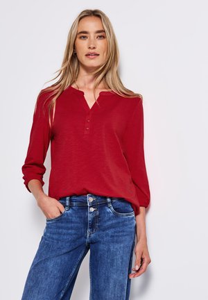 Haut rouge à manches longues avec un col en V et trois boutons, associé à un jean en denim bleu. Le tissu semble doux avec une coupe décontractée.