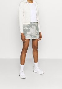 Chaqueta blanca con cremallera, ajuste corto; camiseta blanca; falda mini gris con camuflaje y bolsillo frontal; zapatillas blancas con toques negros; calcetines hasta el tobillo.