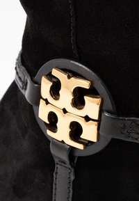 Botín de ante negro que presenta un acento de hardware en tono dorado con un distintivo logo de doble "T", asegurado por una correa de cuero negro.