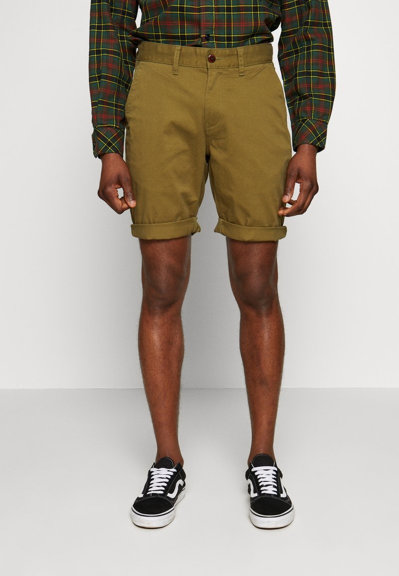 Homme portant un short kaki retroussé, des baskets noires et blanches, et une chemise à manches longues à carreaux verts et rouges, debout devant un fond neutre.