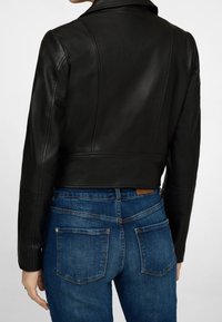 Chaqueta de cuero negra recortada con costuras de hombro pronunciadas y cuello con muesca, combinada con jeans de mezclilla azules que tienen bolsillos traseros.