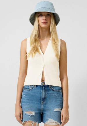 Junge Frau trägt eine ärmellose cremefarbene Weste mit Knöpfen, abgenutzte blaue Jeansshorts und einen hellblauen Fischerhut vor einfarbigem Hintergrund.