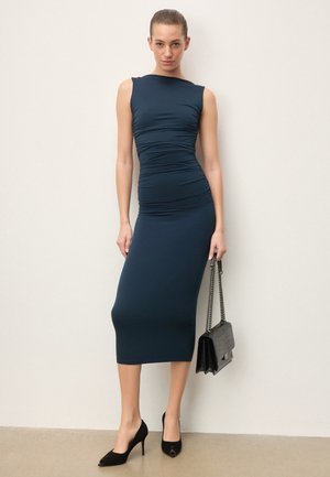 Femeie purtând o rochie midi bleumarin fără mâneci, cu pliuri, pantofi cu toc ascuțiți negri, ținând o geantă neagră cu lanț, stând în picioare în fața unui perete simplu.