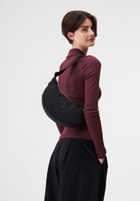 Schwarze, geschwungene Tasche, die über der Schulter getragen wird, aus strukturiertem Stoff, mit roten runden Akzenten und einem breiten, verstellbaren Riemen.