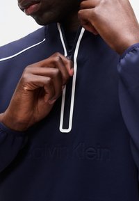 Άτομο που κλείνει το φερμουάρ στον γιακά ενός μπλε σκούρου μπουφάν Calvin Klein με λευκή επένδυση σε φερμουάρ και ώμο.