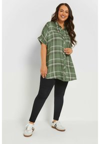 Camisa a cuadros verde de botones con mangas cortas, combinada con leggins negros y zapatillas blancas, de pie frente a un fondo liso.