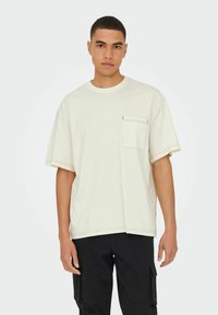 Cremefarvet oversized t-shirt lavet af bomuld, med en brystlomme og kontrastfarvet sort syning, stylede med sorte cargo bukser.