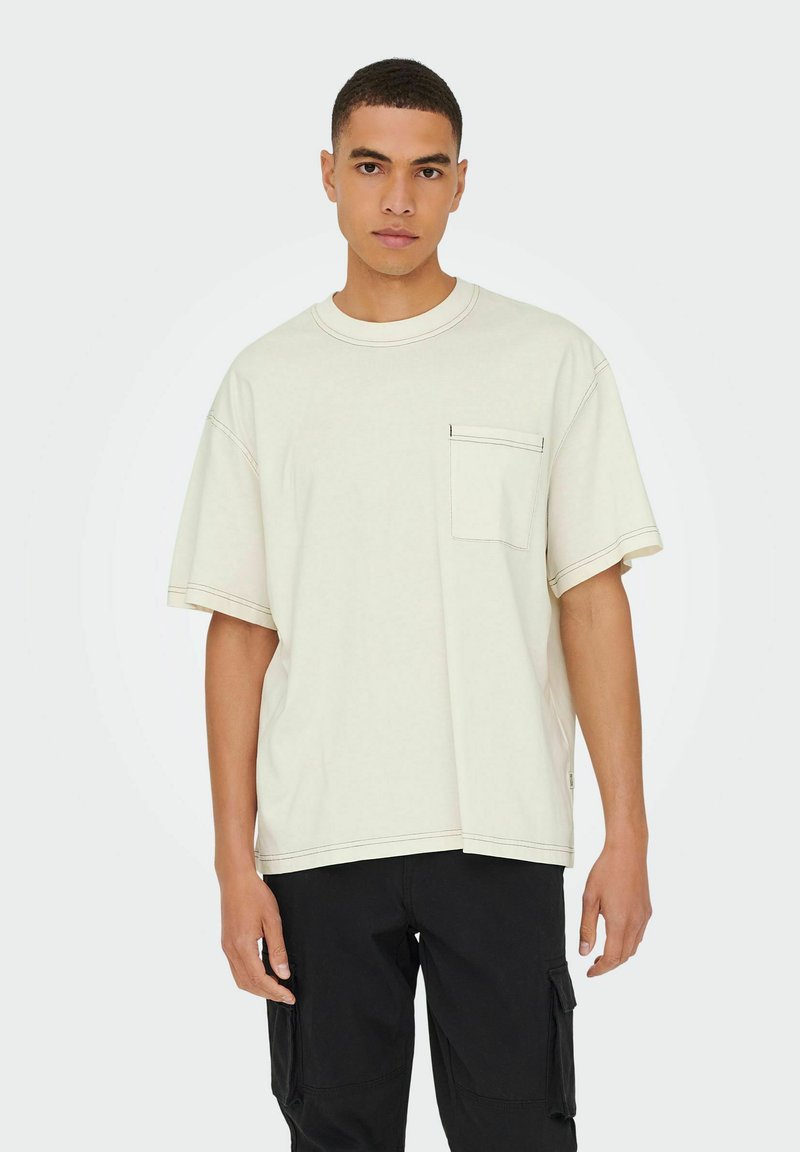 Cremefarvet oversized t-shirt lavet af bomuld, med en brystlomme og kontrastfarvet sort syning, stylede med sorte cargo bukser.