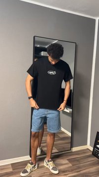 Jeune homme portant un t-shirt noir avec un motif, un short en jean et des baskets, se tient devant un miroir en pied dans une pièce aux murs gris et au sol en bois.