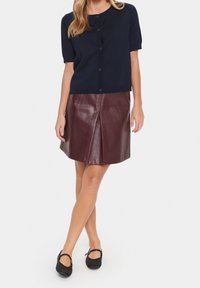 Cardigan bleu marine à manches courtes avec fermeture à boutons, associé à une jupe en cuir bordeaux à plis. Chaussures à brides noires complètent le look.