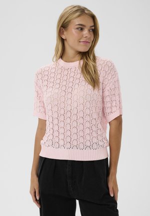 Femme portant un pull en maille manches courtes rose clair avec un motif en dentelle et un pantalon noir taille haute, regardant vers sa droite.