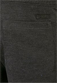 Zwarte katoenstof met een achterzak met zichtbare stiksels en een genaaide label. De textuur lijkt glad en iets pluizig.