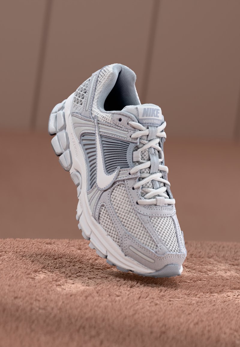 Jasnoszare buty biegowe Nike z siateczkową cholewką, wyściełanym językiem oraz grubą, teksturowaną podeszwą z żelowymi amortyzatorami. Sznurówki są płaskie i splątane.