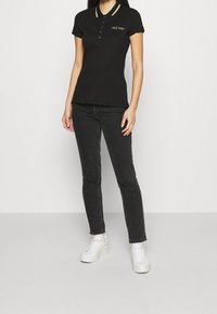 Sort polo skjorte med korte ærmer, guldstribede krave og logo detalje. Kombinationen er med sorte jeans og hvide sneakers.