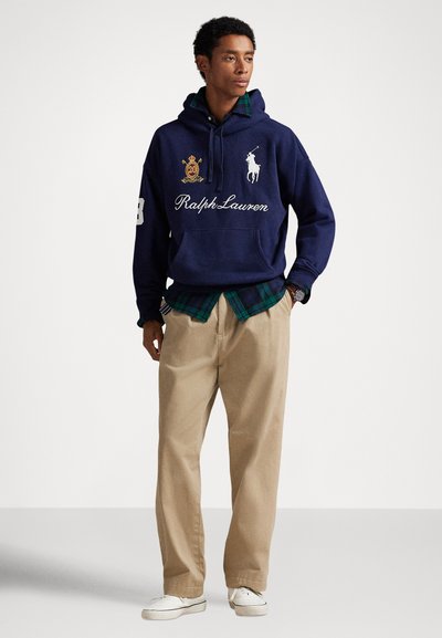 Νέος άντρας που φοράει μπλε σκούρο φούτερ Ralph Lauren, πράσινο πουκάμισο καρό, μπεζ παντελόνι, λευκά αθλητικά παπούτσια και ρολόι χειρός, στέκεται μπροστά σε απλό φόντο.