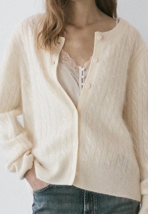 Femme portant un cardigan crème en maille torsadée sur un caraco blanc bordé de dentelle avec un jean en denim bleu, main dans la poche.