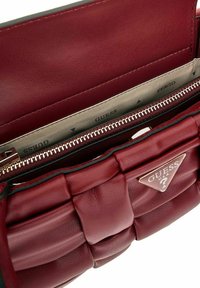 Interno della borsa in pelle bordeaux con texture trapuntata, scomparto con zip e fodera stampata con il marchio "GUESS". Finiture in tonalità oro.