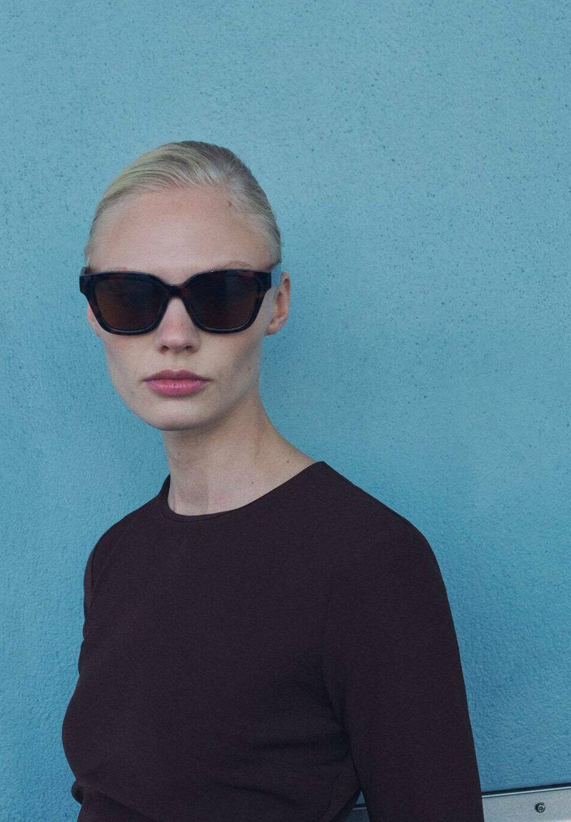 Čierne oversized slnečné okuliare s matným povrchom, v kombinácii s tmavohnedým priliehavým tričkom s dlhým rukávom na svetlomodrom pozadí.