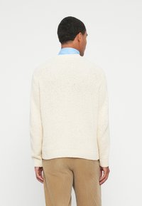 Homem com um suéter de malha creme, camisa de colarinho azul claro e calças de veludo cotelê cor claras, de pé de frente para um fundo branco.