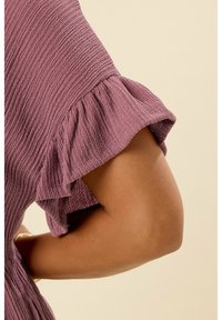 Tessuto viola testurizzato con dettagli di maniche arricciate, caratterizzato da un sottile motivo a coste. Primo piano di un braccio su uno sfondo chiaro.