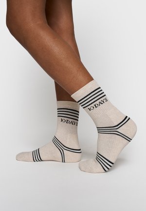 Jambes portant des chaussettes beiges mi-mollet avec des rayures noires et le texte "10 DAYS", un pied à plat et l'autre sur la pointe des pieds, sur un fond uni.