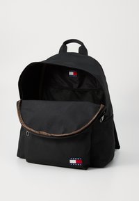 Zaino nero con un cappuccio arrotondato, rifiniture marroni e una toppa con logo sul davanti. Presenta una tasca in rete all'interno e due spallacci.