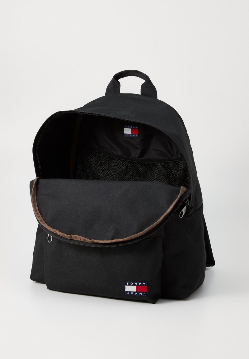 Schwarzer Rucksack mit einer runden Klappe, braunen Akzenten und einem Logo-Patch auf der Vorderseite. Verfügt über eine Netzinnentasche und zwei Schultergurte.