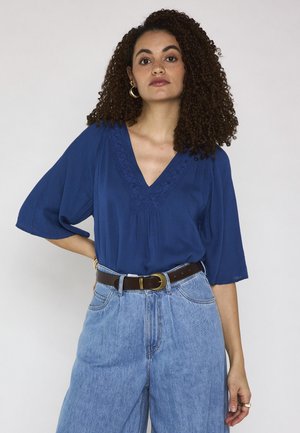 Donna con capelli ricci in piedi con la mano sui fianchi, indossa una camicetta blu navy con scollo a V e jeans azzurri a vita alta con cintura marrone.