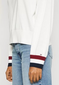 Weißer Pullover mit gerippten Bündchen in Marineblau, Rot und Weiß gestreift. Wird mit hellblauen Jeans getragen und zeigt weichen Stoff sowie eine entspannte Passform.