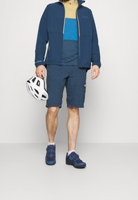 Marinblå vattentät jacka med dragkedja, lagd över en blå och beige färgblockerad skjorta. Marinblå shorts, blå vandringsskor och en vit hjälm.