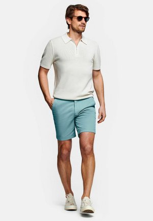 Man draagt een zonnebril, wit T-shirt met korte mouwen en ritskraag, turquoise korte broek en witte sneakers, lopend met één hand in de zak.