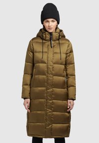 Manteau matelassé long vert olive avec des coutures horizontales, une capuche, des cordons de serrage noirs et des poches latérales. Porté avec un bonnet en tricot noir.