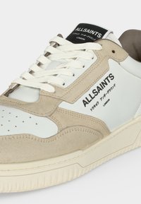 Baskets AllSaints blanches et beiges avec lacets blancs et texte de logo noir sur le côté et la languette indiquant AllSaints London avec des caractères japonais.