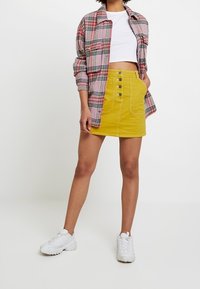 Jupe en denim jaune courte avec des boutons sur le devant, associée à une chemise en plaid rouge et noir, et des baskets blanches.