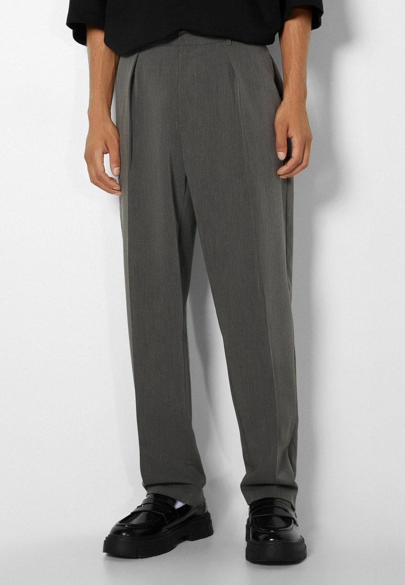 Man som bär lösa grå skräddarsydda byxor och svarta kraftiga loafer-skor, stående mot en vit bakgrund.